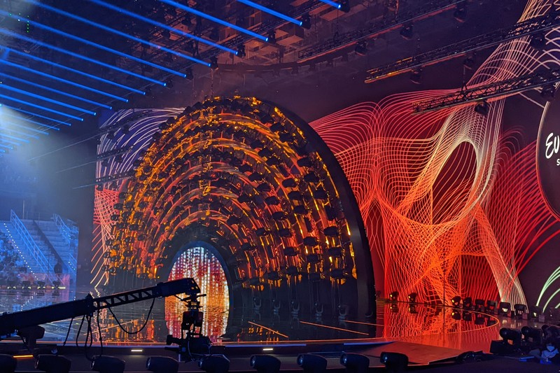 The elaborately lit stage of the Eurovision Song Contest with dramatic orange and blue lighting, camera crane, and audience silhouettes