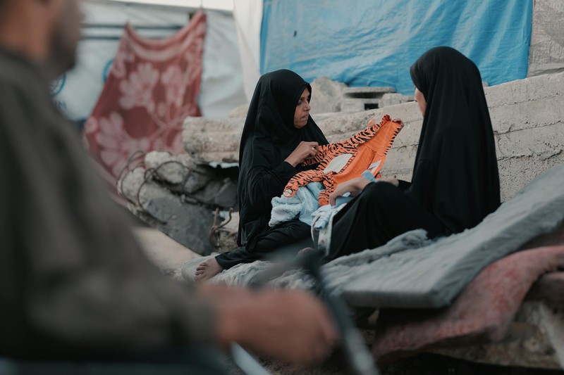 Two women in hijabs sit among rubble and makeshift shelters in Gaza