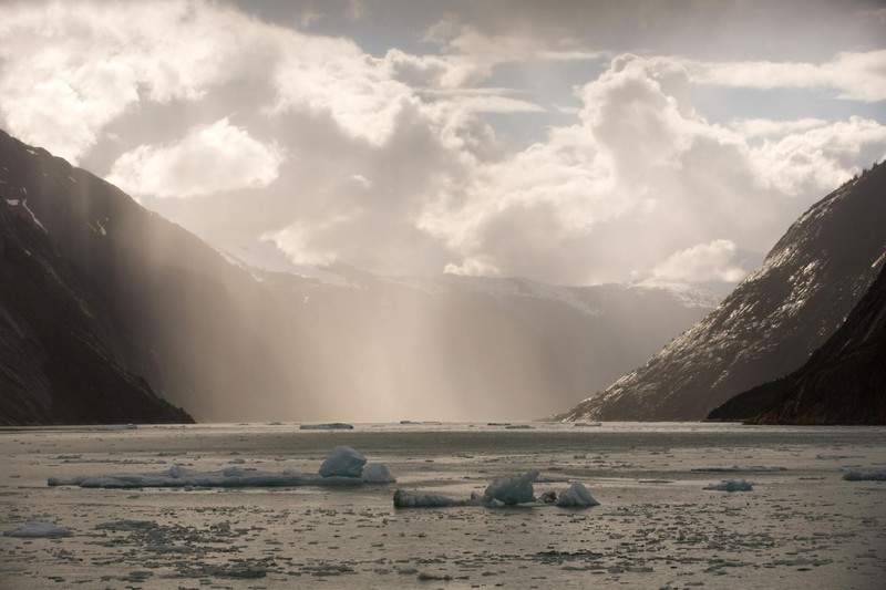 Ice floes float in a glacial Alaskan fjord with steep mountains and dramatic sunbeams breaking through overcast skies