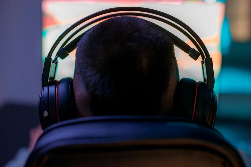 A gamer wearing headphones intently playing a video game on a computer in a dark room