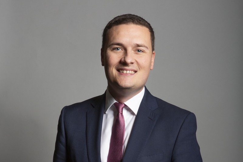 Official portrait of Wes Streeting MP wearing a navy suit and burgundy tie against a grey background