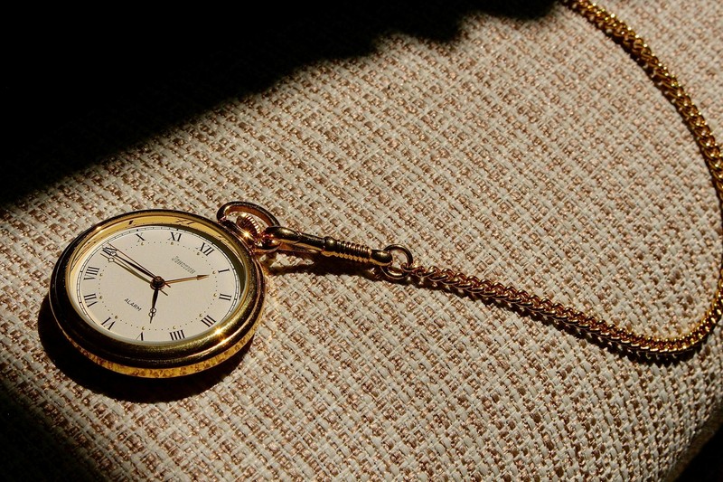 A vintage gold pocket watch with chain resting on woven fabric in warm sunlight