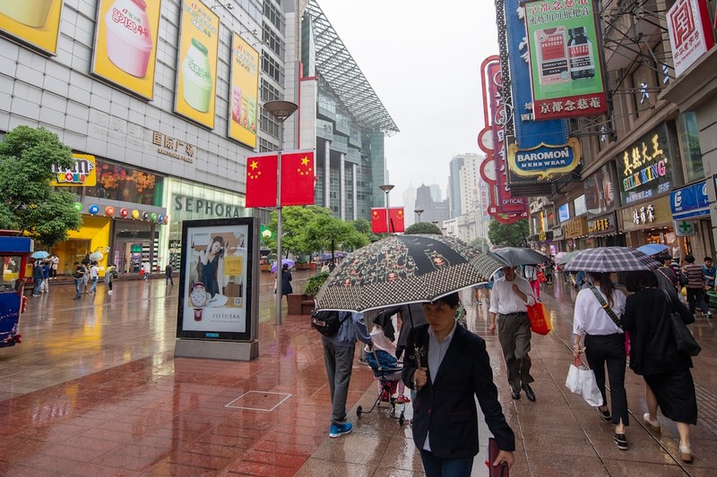 Shoppers walk with umbrellas on a rainy commercial street lined with neon signs and Chinese national flags in Shanghai.