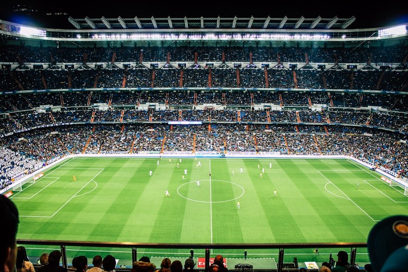 A packed football stadium during a match, viewed from an elevated angle with players on the pitch and fans filling the stands