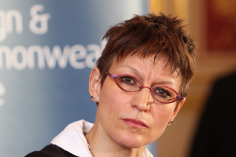 A woman with short brown hair and rectangular glasses at a formal indoor event with a blue backdrop