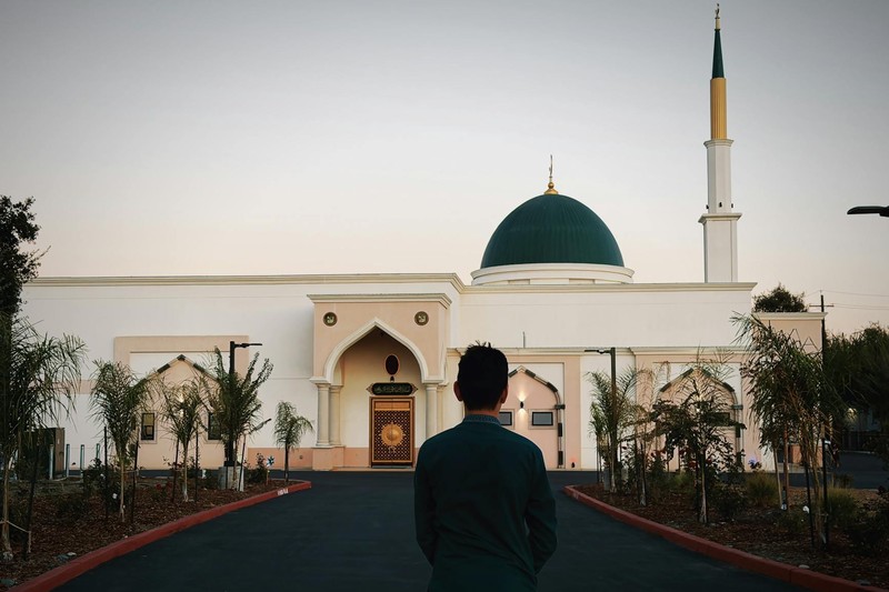 A mosque with a green dome and minaret at dusk, with a person standing on the pathway facing the entrance
