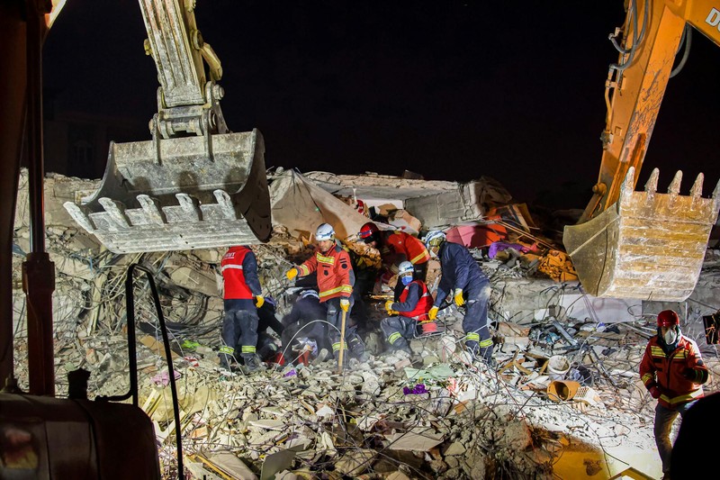 Nighttime rescue operation at a collapsed building site after an earthquake, with emergency workers and excavators clearing debris