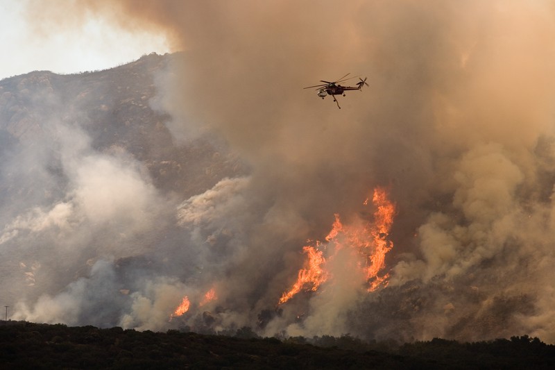 A helicopter drops water and fire retardant on a raging wildfire burning across a hillside, with thick smoke and bright flames visible across the mountainside.