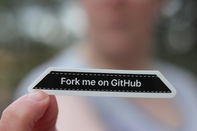 A hand holding a black sticker with white text reading 'Fork me on GitHub'