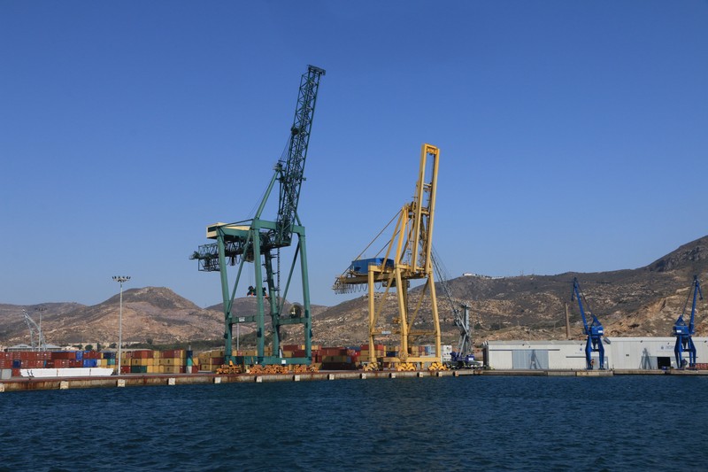 Port cranes and stacked colorful shipping containers at a harbor with mountains in the background