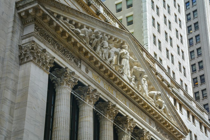 The neoclassical facade of the New York Stock Exchange building with columns and sculptured pediment set against modern skyscrapers