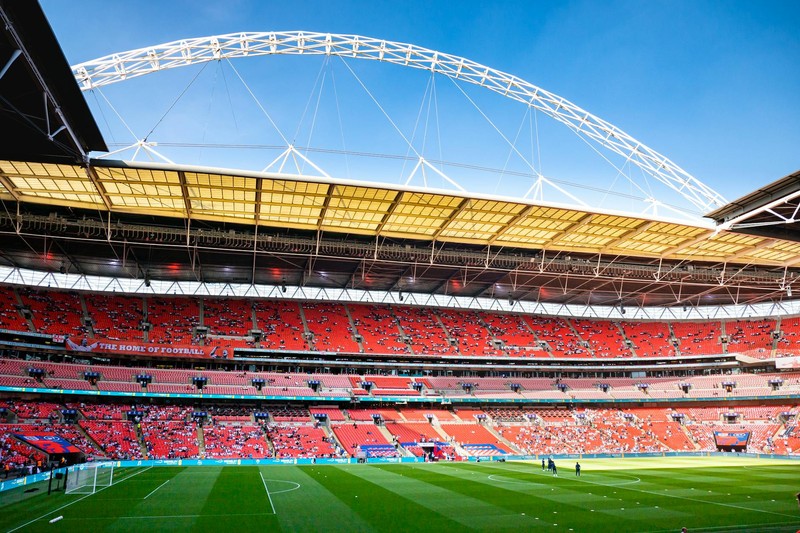 Wembley Stadium in London under clear blue skies, viewed from the pitch with its iconic arch and red seating tiers
