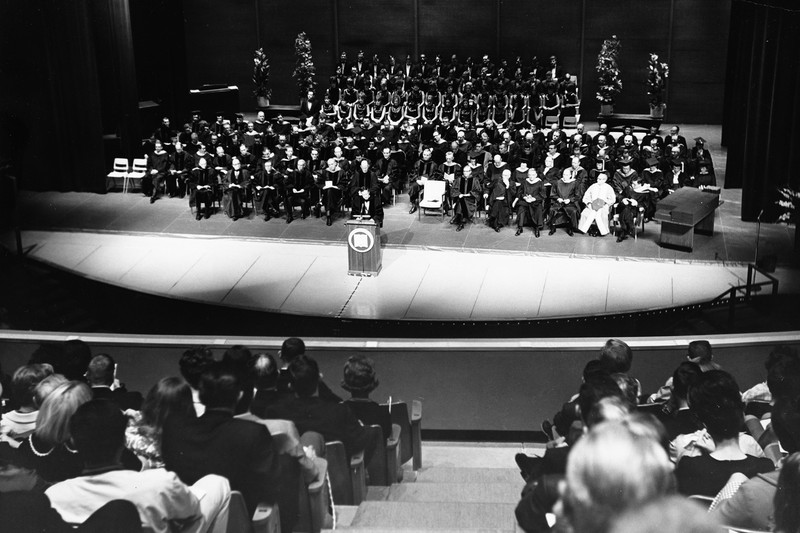 A formal ceremony in a large auditorium with rows of people on stage and an audience seated in the foreground