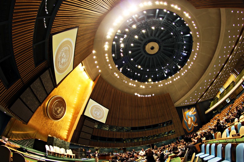 The United Nations General Assembly Hall in New York, viewed from a low angle showing the domed ceiling, UN emblems, and rows of delegates' seats.