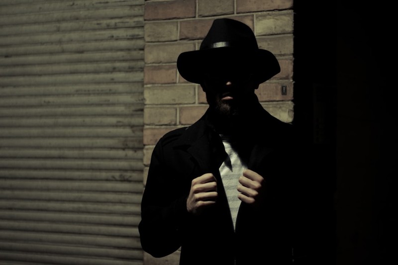A man in a fedora and dark coat stands in a dimly lit urban alleyway, partially obscured by shadow