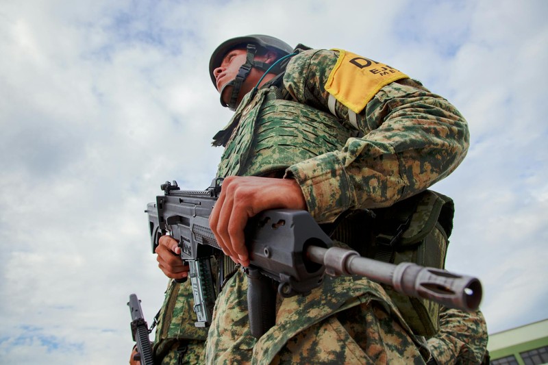 A soldier in camouflage uniform holds a rifle under a blue sky