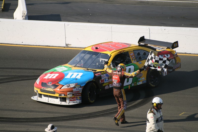 Kyle Busch celebrates with a checkered flag beside his #18 M&M's Toyota Camry after a NASCAR victory at Infineon Raceway