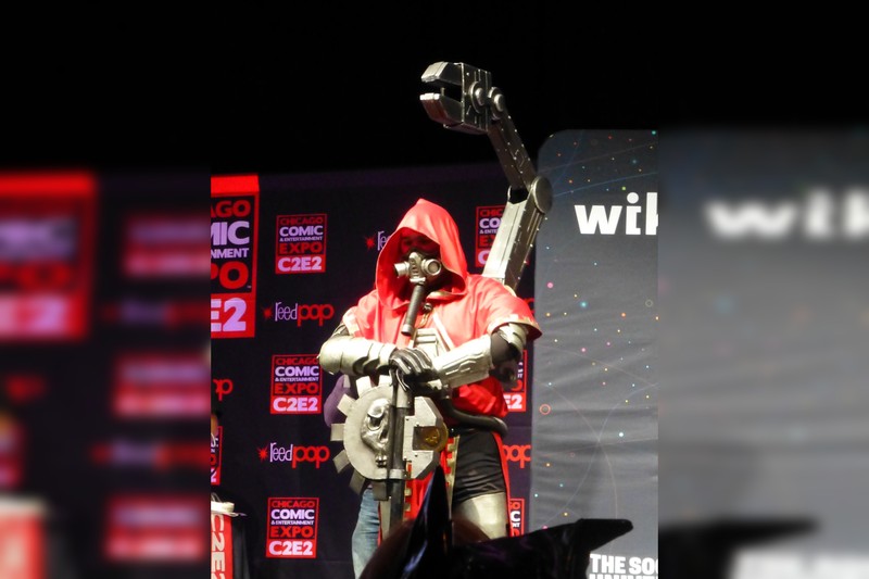 A cosplayer dressed as a Warhammer 40K Tech-Priest in a red hooded robe with a mechanical arm prop at C2E2 2015