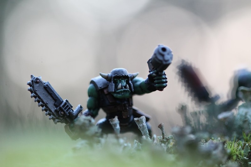 A Warhammer 40K Ork miniature figurine wielding a melee weapon and pistol on a tabletop battlefield diorama