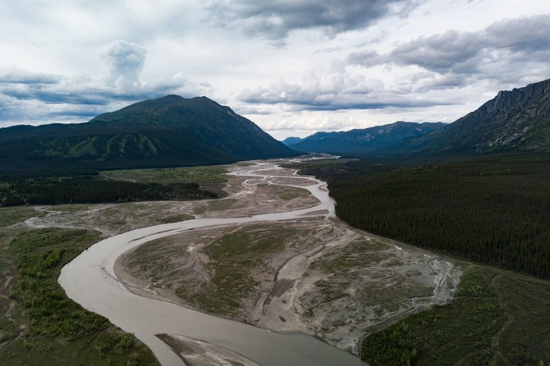 Aerial view of a braided, sediment-laden river winding through a vast Yukon valley between forested mountains under overcast skies