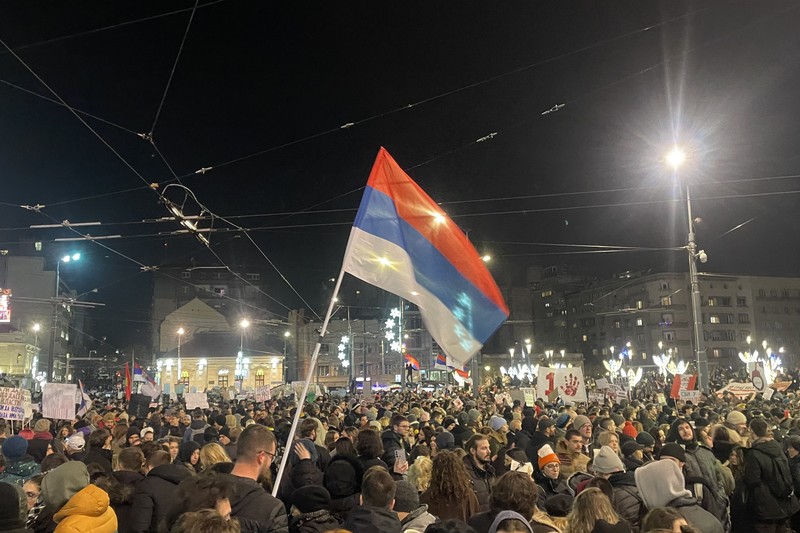 A large crowd of protesters fills a Belgrade city square at night, with Serbian flags waving under streetlights