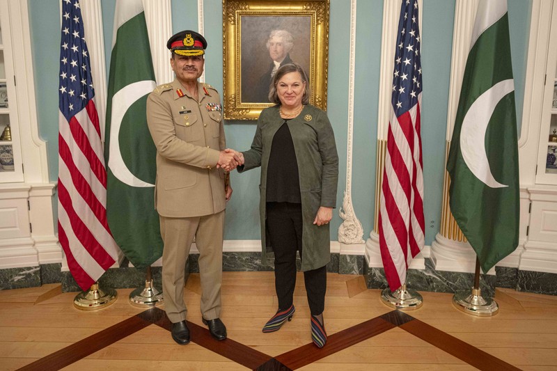 A handshake between a Pakistani military officer and a U.S. official with American and Pakistani flags in the background