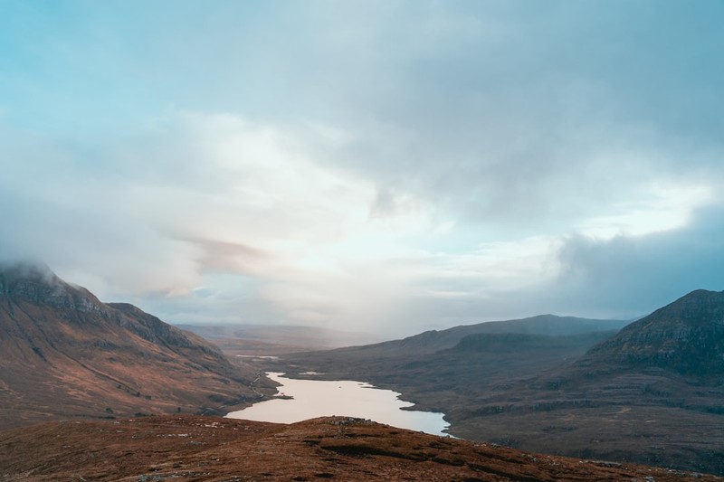 A narrow loch nestled between rugged, dry brown mountains in the Scottish Highlands under an overcast sky