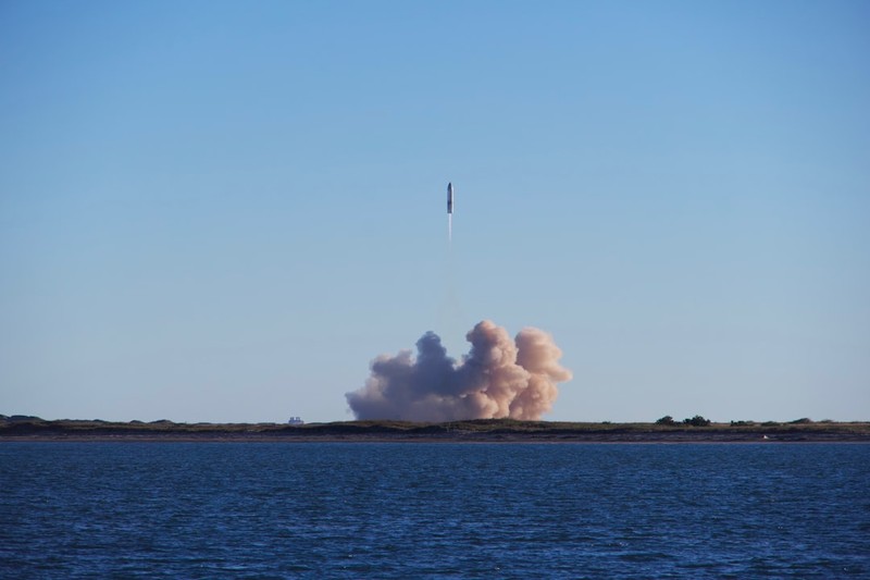 A SpaceX Starship rocket lifts off from a coastal Texas launch site, ascending into a clear blue sky with a massive plume of smoke billowing below.