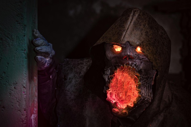A menacing hooded monster with glowing orange eyes and a fiery mouth emerges from a dark, grimy corner in detailed horror cosplay