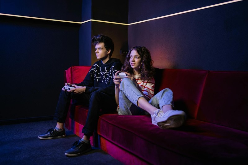 Two people sitting on a red couch playing video games with controllers in a dimly lit room