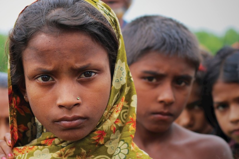 A young Bangladeshi girl in a floral headscarf gazes solemnly at the camera, with other children visible behind her in an outdoor setting.