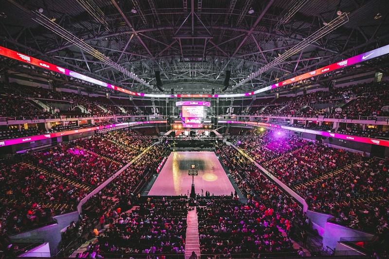 A packed indoor sports arena with a basketball court illuminated under vibrant purple and pink lighting
