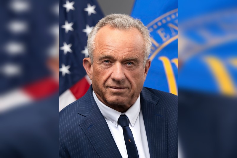 Official portrait of Health and Human Services Secretary Robert F. Kennedy Jr., wearing a dark suit and tie, with American and HHS flags in the background