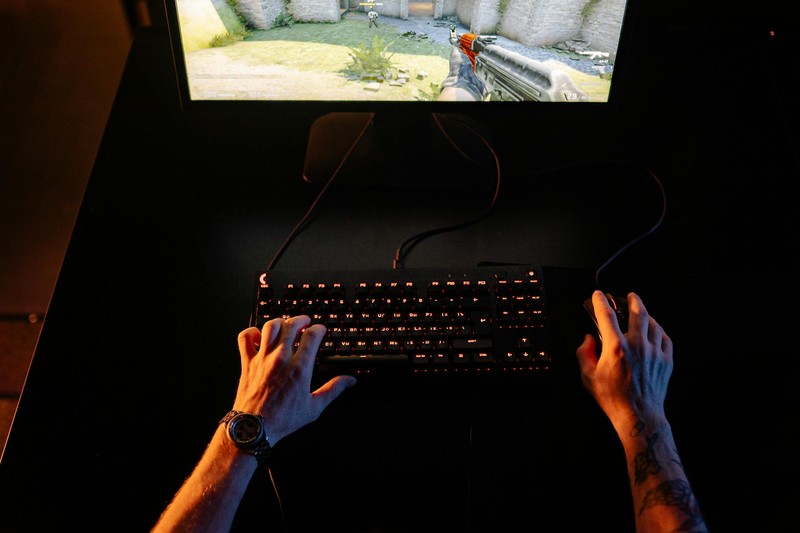 A gamer's hands on a backlit keyboard and mouse while playing a first-person shooter game on a monitor in a dark room