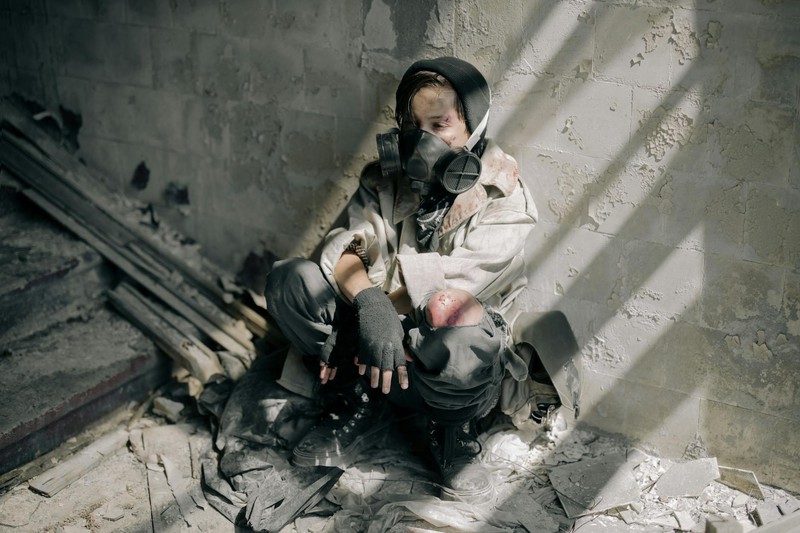 A person wearing a gas mask sits in a dilapidated, debris-filled abandoned building with harsh shadows cast across the walls
