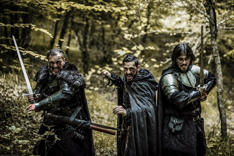 Three medieval warriors armed with swords stand in a dense autumn forest
