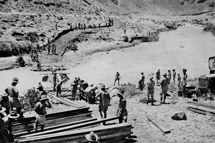 Historical black-and-white photograph of soldiers bridging the Litani River near Merjayun, with workers and construction materials along a rocky riverbed amid arid hills.