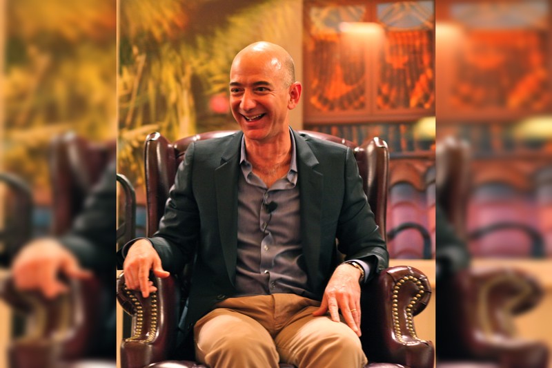 Jeff Bezos seated in a dark armchair during a media appearance, wearing a blazer and speaking into a clip-on microphone.