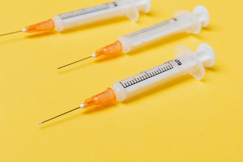 Three plastic medical syringes with orange needle hubs arranged on a bright yellow surface
