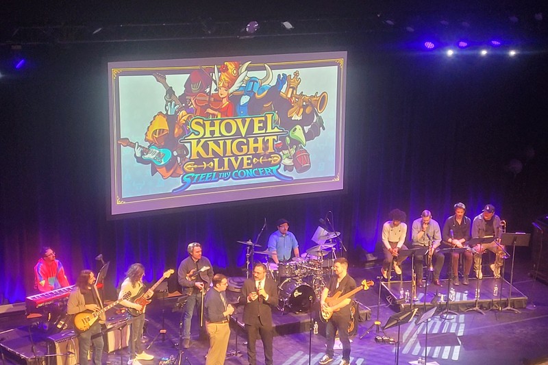 A live Shovel Knight concert performance on stage with musicians arranged in a band setup, a large screen displaying 'Shovel Knight Live!!' with pixel-art characters behind them.