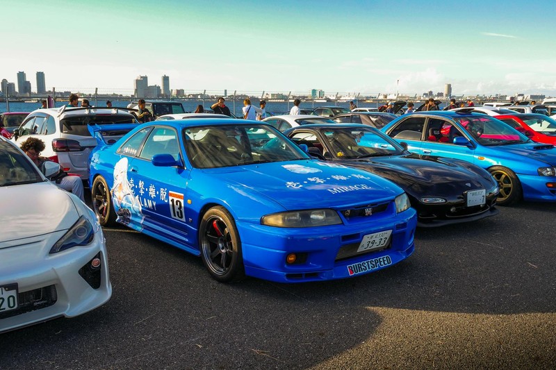 A blue Nissan Skyline R34 at an outdoor car meet surrounded by other modified sports cars