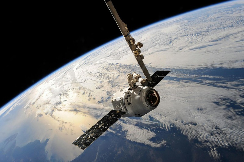A satellite with extended solar panels orbits above Earth, with dramatic cloud formations and the curvature of the planet visible below against the blackness of space.