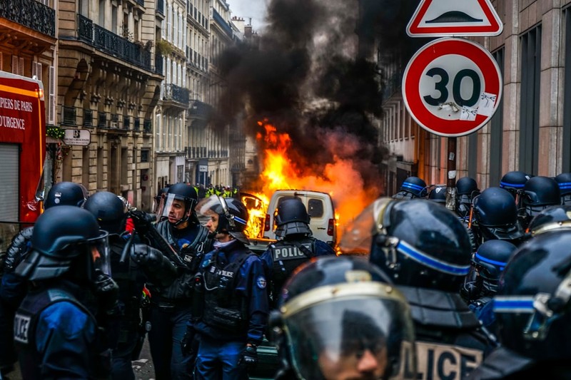 Riot police in tactical gear responding to a burning vehicle on a narrow Paris street with thick smoke billowing