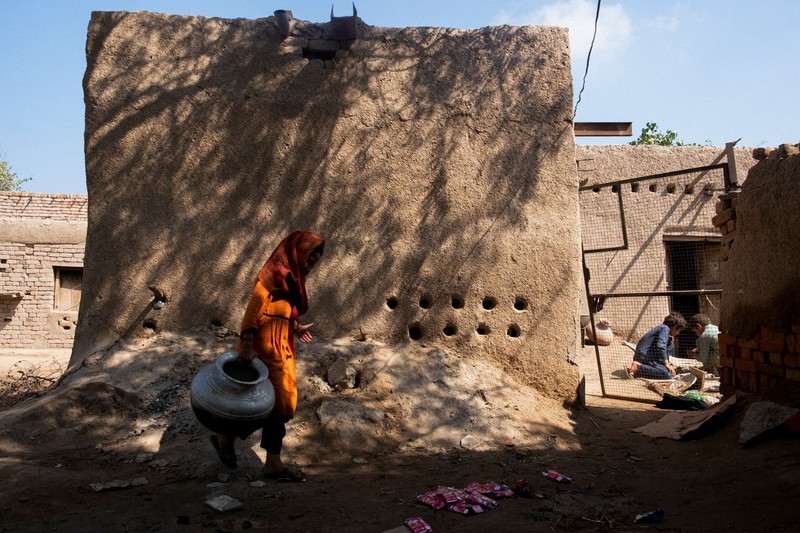 A woman in an orange garment carries a water pot through a sun-dried Pakistani village with traditional mud-brick houses