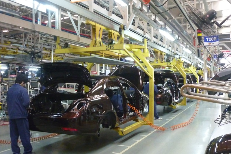 Cars on a yellow assembly line at a Geely factory in Ningbo, China, with workers stationed along the production floor.