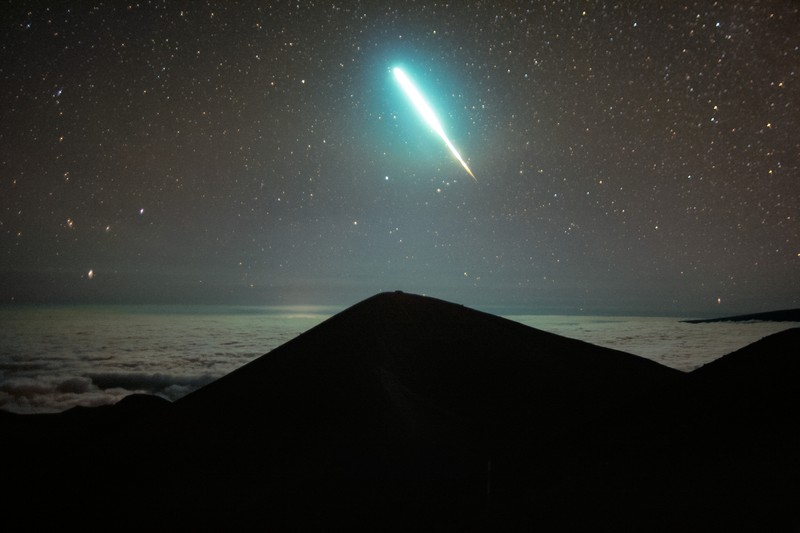 A bright turquoise meteor streaks across a star-filled night sky above a dark mountain silhouette