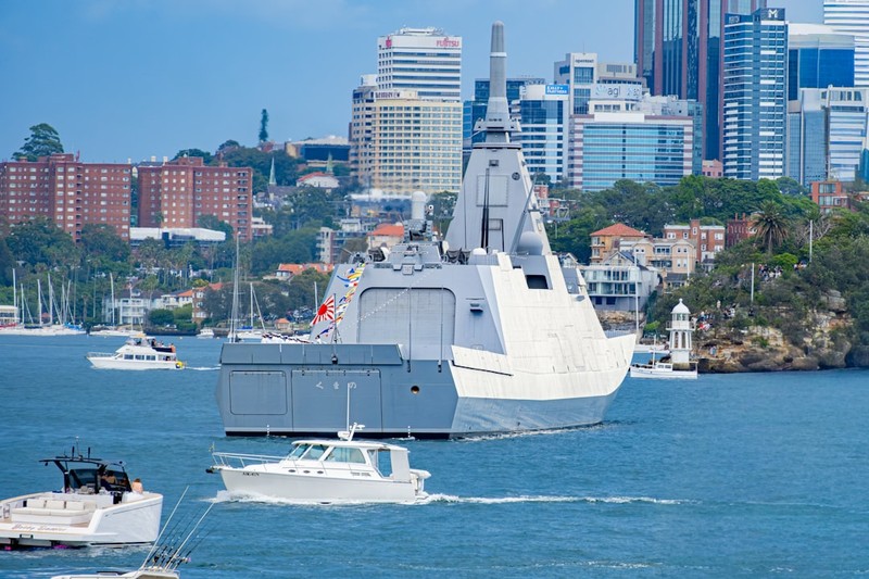 A Mogami-class frigate of the Japan Maritime Self-Defense Force sailing through a harbor with city skyline in the background