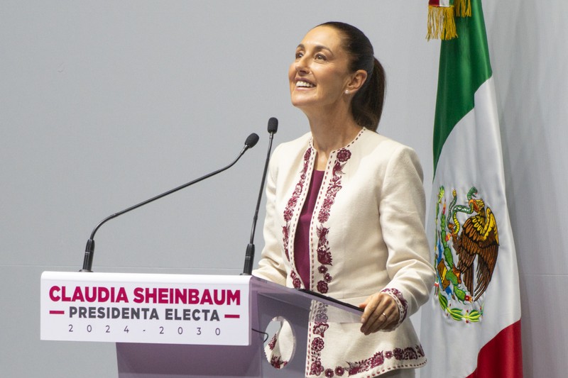 Claudia Sheinbaum stands at a podium labeled Presidenta Electa 2024-2030 with the Mexican flag beside her