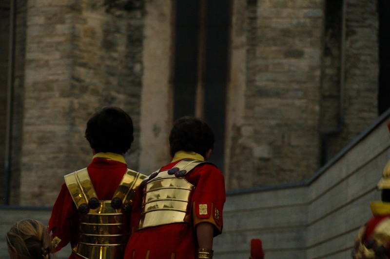 Two figures in Roman-style red and gold armor approaching an ancient stone building