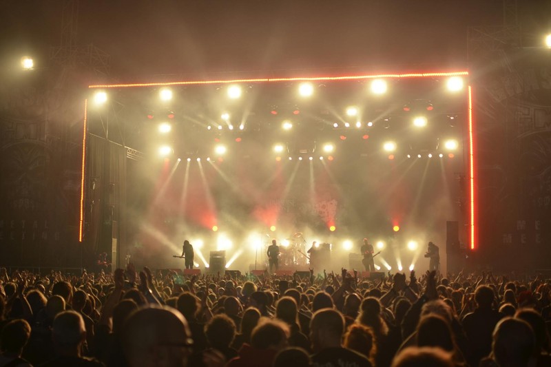A band performs on a brightly lit stage at a live concert with an enthusiastic crowd raising their hands